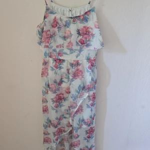 Blommig långklänning med volang - Superfin långklänning med tunna axelband och volang upptill. Klänningen är vit med blommigt mönster i rosa och blå toner, perfekt för sommarens varma dagar. Lätt och luftig med elastisk midja för skön passform.