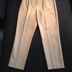 Beiga kostymbyxor med bälte Gina Tricot - Stilrena beiga kostymbyxor från Gina Tricot i storlek 38. Byxorna har hög midja, raka ben och medföljande bälte i samma tyg. Tillverkade i ett mjukt, vävt material med dragkedja och knäppning framtill. Perfekta för en clean och dressad look.