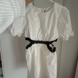 Vit kortklänning med svart knytband - Superfin vit kortklänning från H&M med puffärmar och svart knytband i midjan. Klänningen har rund halsringning och är tillverkad i ett lätt bomullstyg. Perfekt för dig som gillar en stilren look med en twist!