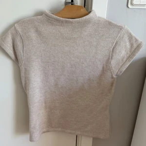 Beige stickad polotopp - En enkel och stilren beige stickad polotopp med korta ärmar. Toppen har en mjuk och skön känsla, perfekt för att matcha med jeans eller kjol. Den har en högre halsringning som ger en clean look.