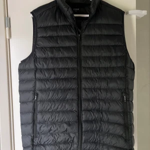 Svart dunväst från Everest - Säljer en svart dunväst från Everest i storlek L. Västen har quiltad design, hög krage och dragkedja framtill samt två fickor med dragkedja. Perfekt att slänga över en hoodie för extra värme. Enkel och stilren look. Inga tecken på användning. Passar M