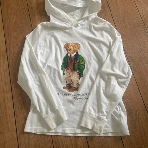 Vit hoodie Polo Bear från Ralph Lauren - Vit hoodie från Polo Ralph Lauren med ikoniska Polo Bear-trycket framtill. Tröjan har huva, långa ärmar och är tillverkad i 100% bomull för en skön och avslappnad känsla. Perfekt för sommarn