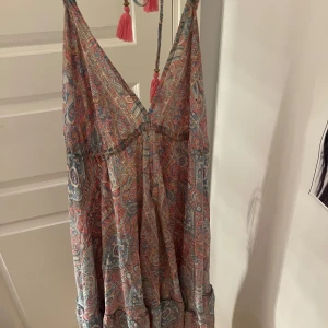 Paisleymönstrad långklänning med tofsar - Superfin långklänning med paisleymönster i rosa, blått och beige. Djup v-ringning, smala axelband med rosa tofsar och avslappnad passform. Perfekt för varma dagar och ger en bohemisk vibe. Köpt utomlands för ca 350kr 