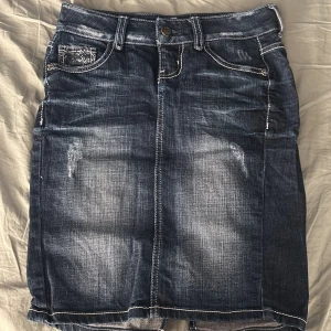 Mörk jeanskjol från Guess med bling - Snygg mörkblå jeanskjol från Guess med slitna detaljer och bling på bakfickan.  Den är bara använd nån gång och är förliten för mig. Väldigt liten midja och går ungefär till knäna. Lite slitage men det är inte så märkvärdigt med tanke på stilen