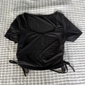 Svart croppad topp med knyt från SHEIN - Svart croppad topp från SHEIN i storlek XS. Toppen har korta ärmar, djup v-ringning och snygga knytband i sidorna för en trendig look. Perfekt att matcha med höga jeans eller kjol för en cool stil.