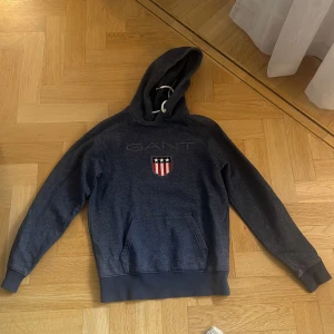Mörkblå hoodie från GANT XS - Snygg mörkblå hoodie från GANT med broderad logga och sköld på bröstet. Tröjan har huva med vit snörning och en stor magficka. Perfekt för dig som gillar klassisk och sportig stil.