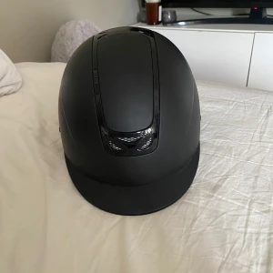 Svart ridhjälm JH Collection - Snygg svart ridhjälm från JH Collection med matt finish och ventilerande detaljer i blankt svart. Hjälmen har justerbart hakband och bekväm insida med mips. 