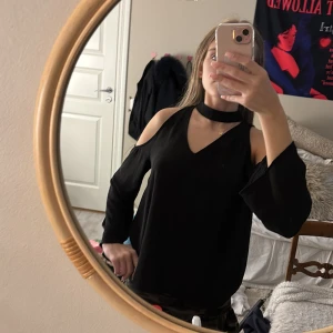 Svart blus med cut out från Gina Tricot - Svart blus från Gina Tricot med v-ringning, långa ärmar och trendiga cut out-detaljer vid axlarna. Snygg chokerkrage och lätt, flowigt tyg som ger en cool look. Perfekt för dig som gillar stilrena och moderna plagg.