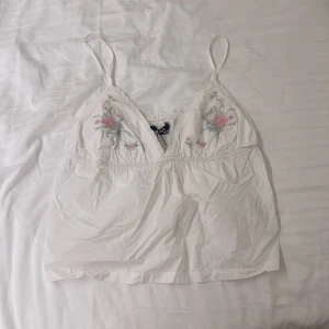 Vit blommig linnetopp från BikBok - Supersöt vit linnetopp från BikBok med tunna axelband och broderade blommor i rosa och grönt på bysten. Toppen har spetsdetaljer längs kanterna och en lätt rynkad resår under bysten för en feminin look. Perfekt för sommarens alla tillfällen! Säljer pga att den inte används. Passar perfekt på mig som är storlek M i vanliga fall.
