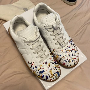 Maison Margiela sneakers med färgstänk - OBS skriv till mig innan ni köper Unika vita sneakers från Maison Margiela med färgstänk i blått, rött, gult och svart över tån. Skorna har klassisk siluett, snörning och gummisula i brunt. Tillverkade i skinn och har en riktigt cool och konstnärlig vibe. 