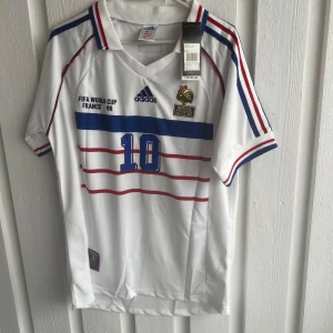Frankrike Zidane 10 VM 98 Adidas tröja - Klassisk vit fotbollströja från Adidas, Frankrikes landslag VM 1998 med Zidane och nummer 10. Tröjan har blå och röda detaljer, broderade märken och tryck, samt ränder på ärmar och bröst. Materialet är lätt och andas, perfekt för fotbollsfans.