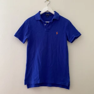 Blå Polo Ralph Lauren t-shirt slim fit - Snygg blå t-shirt från Polo Ralph Lauren i slim fit-modell. Klassisk pikékrage, korta ärmar och orange broderad logga på bröstet. Tillverkad i mjuk bomull som känns skön mot huden. Perfekt för dig som gillar stilren och sportig look.