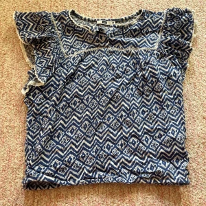 Blåmönstrad topp från WE - Säljer en blåmönstrad topp från WE med volangdetaljer på ärmarna. Toppen har ett unikt geometriskt mönster i blått och vitt samt knappar framtill. Perfekt för en somrig look!