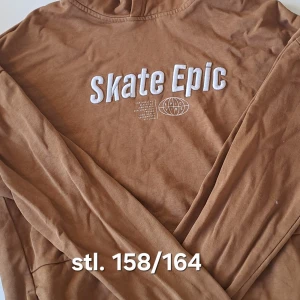 Brun hoodie med Skate Epic-tryck - Säljer en brun hoodie med Skate Epic-tryck på framsidan. Tröjan har långa ärmar och en avslappnad passform, perfekt för skate-entusiaster. Materialet känns mjukt och bekvämt mot huden. Tröjan har luva, men syns knappt på bilden.