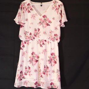 Blommig klänning med v-ringning - Säljer en söt blommig klänning med v-ringning och korta ärmar. Klänningen har ett elastiskt band i midjan för en bekväm passform. Perfekt för sommarens varma dagar!