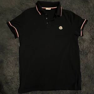 Moncler pikétröja  - Snygg svart pikétröja från Moncler med klassisk krage och knappar. Tröjan har vita och röda detaljer på kragen och ärmsluten samt Moncler-logga på bröstet. 