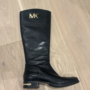 Svarta stövlar från Michael Kors - Snygga svarta stövlar i skinn från Michael Kors med gulddetaljer. Stövlarna har en dragkedja på sidan och en dekorativ MK-logga upptill. Perfekta för en stilren look.