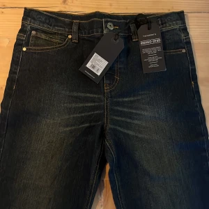 Mörkblå jeans från Lyle & Scott - Snygga mörkblå jeans från Lyle & Scott med klassisk femficksdesign och kontrasterande sömmar. Jeansen har en knappgylf och är tillverkade i ett slitstarkt denimtyg. Perfekta för en stilren look.