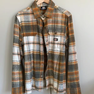 Rutig skjorta från Dickies - Säljer en snygg rutig skjorta från Dickies i orange, grått och vitt. Skjortan har långa ärmar och knappar framtill. Perfekt för en avslappnad stil med en klassisk touch. Passar bra till jeans eller chinos. Har använt den ett antal gånger men skjortan är så gott som ny. Älskade att använda den men behöver rensa ut garderoben.