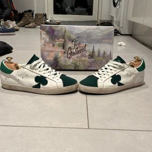 Vita och gröna sneakers från The Gallerist - Snygga vita sneakers med gröna detaljer från The Gallerist. Skorna har en unik design med en grön klöver på sidan och texten 'I My Gallerist' på innersulan. Perfekta för dig som vill ha något annorlunda. Använda ett fåtal gånger!!