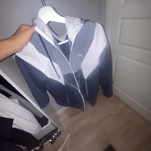 Grå och vit huvtröja med dragkedja - Säljer en snygg huvtröja från BOSS i grått och vitt med dragkedja framtill. Tröjan har en modern design med kontrasterande paneler och en justerbar huva med snören. Perfekt för en avslappnad stil.