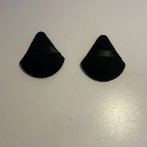 Svarta puderpuffar - Säljer ett par svarta puderpuffar med en slät yta och en diskret glansig remsa. Perfekta för att göra ditt smink bättre. Mjuka och och små så dem får plats i din necessär.