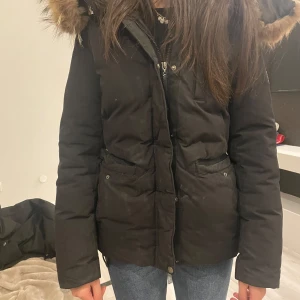 Svart parkajacka med pälsdetaljer - Säljer en svart parkajacka med pälsdetaljer på huvan. Jackan har en dragkedja framtill och långa ärmar. Perfekt för kyliga dagar med sin varma och stilrena design.