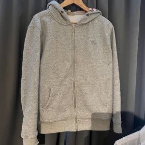 Grå hoodie från Burberry - Säljer en stilren grå hoodie från Burberry med dragkedja och broderad logga på bröstet. Tröjan har en klassisk design med huva och långa ärmar. Perfekt för en avslappnad look.