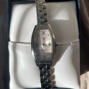 Tommy Hilfiger Klocka - Elegant klocka från Tommy Hilfiger med silverfärgad boett och armband. Urtavlan har romerska siffror och en subtil logotyp. Perfekt för en stilren look. Helt oanvänd nypris ungefär 1000.