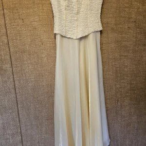 Vit långklänning med rynkad topp - Elegant vit långklänning med rynkad topp och smala axelband. Kjolen är i ett slätt och glänsande material som ger en fin fall. Perfekt för speciella tillfällen.