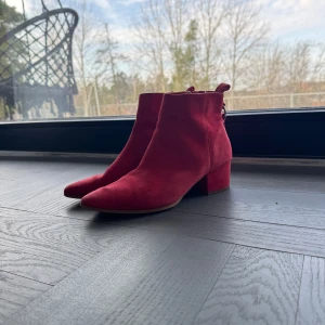 Röda mocka boots Steve madden  - Snygga rödorangea boots i mocka med en låg klack. De har en dragkedja på sidan för enkel på- och avtagning samt en dekorativ rem med spänne baktill. Perfekta för att ge en färgklick till din outfit.