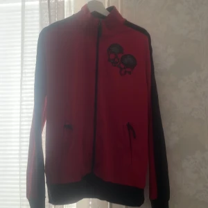 Röd jacka med dödskallemotiv - Säljer en cool röd jacka med svarta ärmar och ett dödskallemotiv på bröstet. Denna tröja är sjukt snygg dock nästan aldrig använd! Den är i storlek L men funkar som storlek M/S också beror på hur man vill styla 