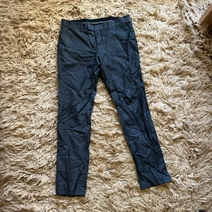 Mörkgråa kostymbyxor från Tige of Sweden - Mörkgråa kostymbyxor med en elegant och stilren design. Perfekta för en formell look. Byxorna har en rak passform och är tillverkade i ett material som ger en fin struktur. Mått: midja-92cm insida ben-89cm