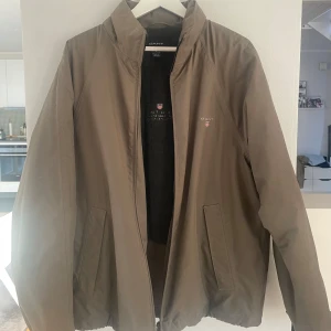 Grön jacka från GANT - Säljer en stilren grön herr jacka från GANT i storlek M. Jackan har en dragkedja framtill och två sidofickor med dragkedja. 