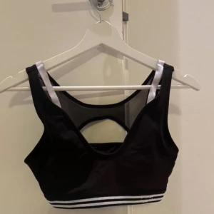 Svart sportsbra från Lacoste - Snygg svart sportbra från Lacoste med vita detaljer och meshpartier. Toppen har en elastisk randig kant i svart och vitt. 