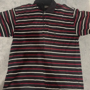 Randig pikétröja från Yves Saint Laurent - Säljer en stilren randig pikétröja från Yves Saint Laurent. Tröjan har svarta, röda och vita ränder och korta ärmar. Perfekt för en avslappnad men ändå elegant look.