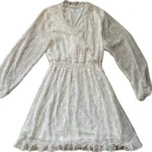 Vit blommig klänning från Vila - Charmig blommig klänning från Vila i ljusa toner. Den har långa ärmar som man kan se igenom, och en smockad midja för en bekväm passform. Volangdetaljer vid halsen och nederkanten ger en romantisk touch. Perfekt för vårens och sommarens tillfällen. Den ser helt ny ut och har endast använts en gång. 