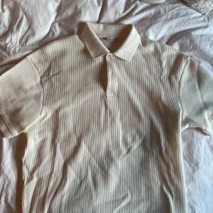 Vit pikétröja från Zara - Säljer en stilren vit pikétröja från Zara. Tröjan har en klassisk krage och knappar framtill. Den är kortärmad och har ett diskret randigt mönster som ger en snygg textur. Perfekt för en avslappnad men ändå uppklädd look. Aldrig använd!