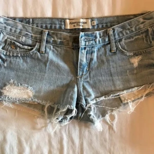 Blå jeansshorts från Abercrombie & Fitch - HAR INTE MIDJEMÅTT!Snygga blå jeansshorts från Abercrombie & Fitch med slitna detaljer och fransar. Perfekta för en avslappnad stil. De har en klassisk femficksdesign och knappgylf.