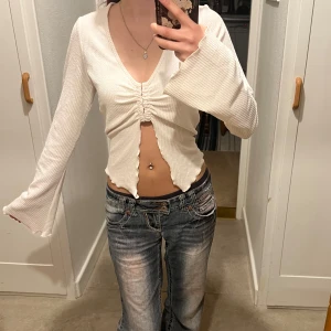 Vit/ beige ribbad topp - Säljer en vit/ beige ribbad topp med v-ringning och långa ärmar. Toppen har en snygg knytdetalj framtill och är croppad med vågig kant. Perfekt för en trendig look!