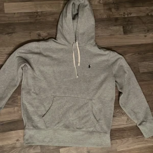 Grå hoodie från Ralph Lauren - Säljer en stilren grå hoodie från Ralph Lauren med en liten broderad logga på bröstet. Den har en klassisk känguruficka och justerbar huva med snören. Perfekt för en avslappnad look.