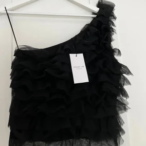 Svart oneshoulder topp från Stockh LM Studio - Snygg svart oneshoulder topp från Stockh LM Studio med volangdetaljer. Perfekt för en kväll ute eller en speciell tillställning. Toppen har en unik design med flera lager av volanger som ger en trendig look.