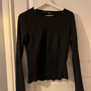 Svart långärmad tröja från 157 - Säljer en svart långärmad tröja från 157. Tröjan har en texturerad yta och en enkel design som passar perfekt till olika outfits. Perfekt för både vardag och speciella tillfällen.