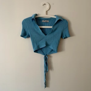 Blå ribbad knytblus - Säljer en snygg blå ribbad blus. Den har en v-ringad design med krage och korta ärmar. Blusen är croppad och har knytband framtill för en trendig look. 