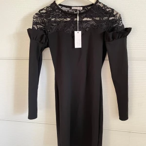 Svart klänning med spetsdetaljer från NLY Trend - Elegant svart klänning från NLY Trend med vackra spetsdetaljer över axlarna och bröstet. Klänningen har långa ärmar och en figurnära passform. Perfekt för en stilfull look.