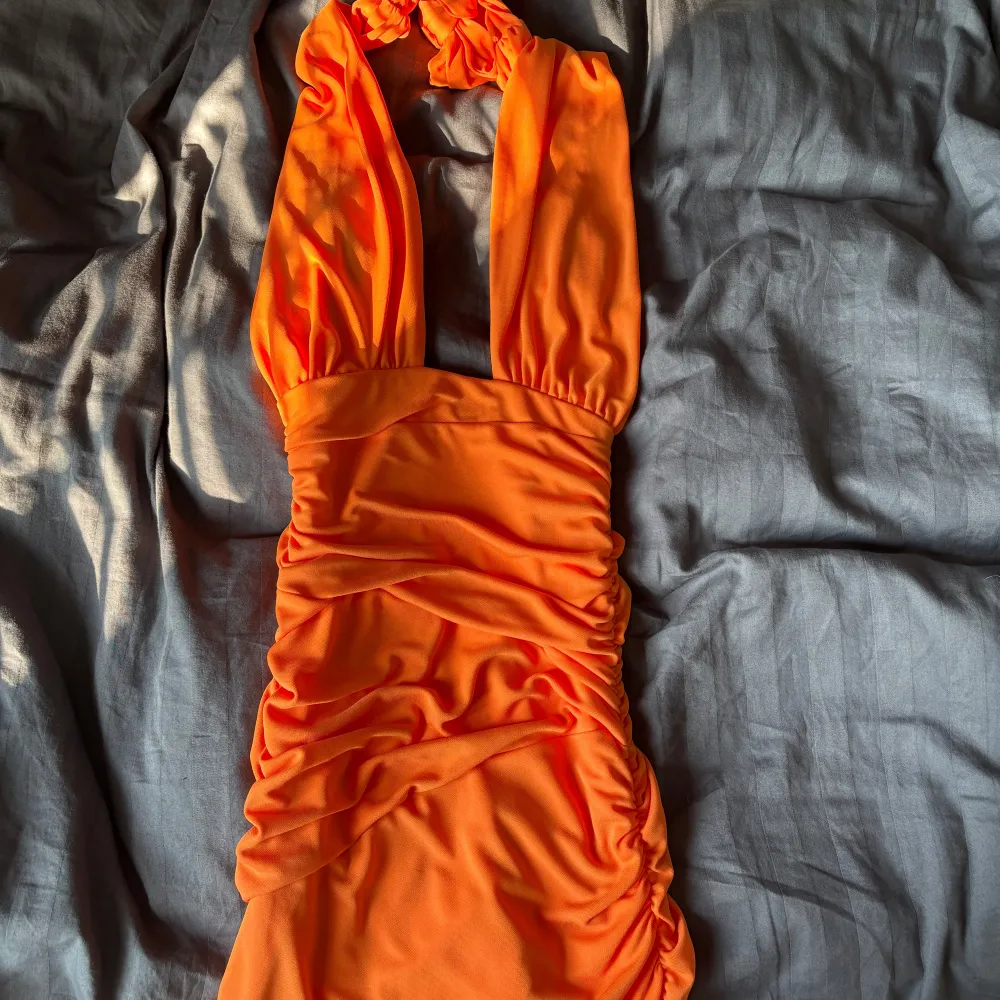 Orange omlottklänning med halterneck och rynkade detaljer. Går att knyta på 2 olika sätt (jmfr bild 1-2) öppen rygg och figurnära. Aldrig använd och normal i storleken.. Mekot.