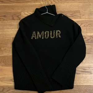 Svart tröja från Zadig & Voltaire - Snygg svart tröja från Zadig & Voltaire med texten 'AMOUR' i glittriga bokstäver på framsidan. Tröjan har långa ärmar och en unik krage med knappar. Perfekt för en stilren look. 