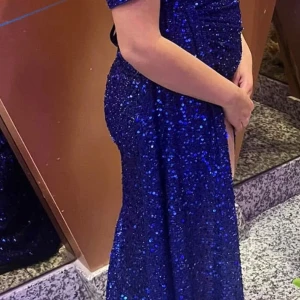 Blå glittrig långklänning - Säljer en fantastisk blå glittrig långklänning med en elegant slits framtill. Klänningen har en tight passform som framhäver figuren och korta ärmar. Perfekt för en glamorös kväll!  