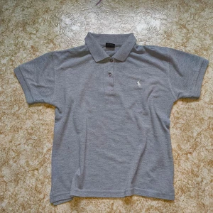 Grå pikétröja från Ralph Lauren - Säljer en klassisk grå pikétröja från Ralph Lauren med korta ärmar och en liten broderad logotyp på bröstet. Passar perfekt för sommaren.