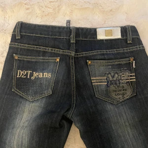 Mörkblå jeans från D2T Jeans - Snygga mörkblå jeans från D2T Jeans, low waist flare med kontrastsömmar och en cool broderad detalj på bakfickan. Jeansen har en klassisk femficksdesign och en knappgylf. Insidan av midjan är fodrad med rött tyg för en extra touch.